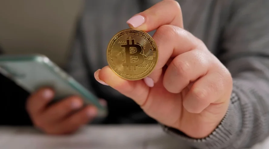 A person holding a golden Bitcoin coin while holding their phone